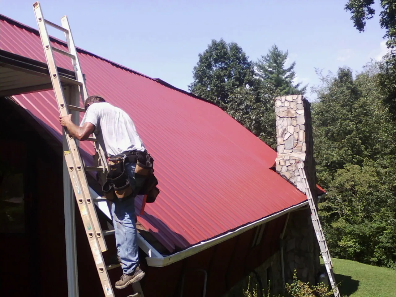 Roofer on a ladder beside a red metal roof and stone chimney — Soffit & Fascia Repair by Roofing Brothers in Bennington